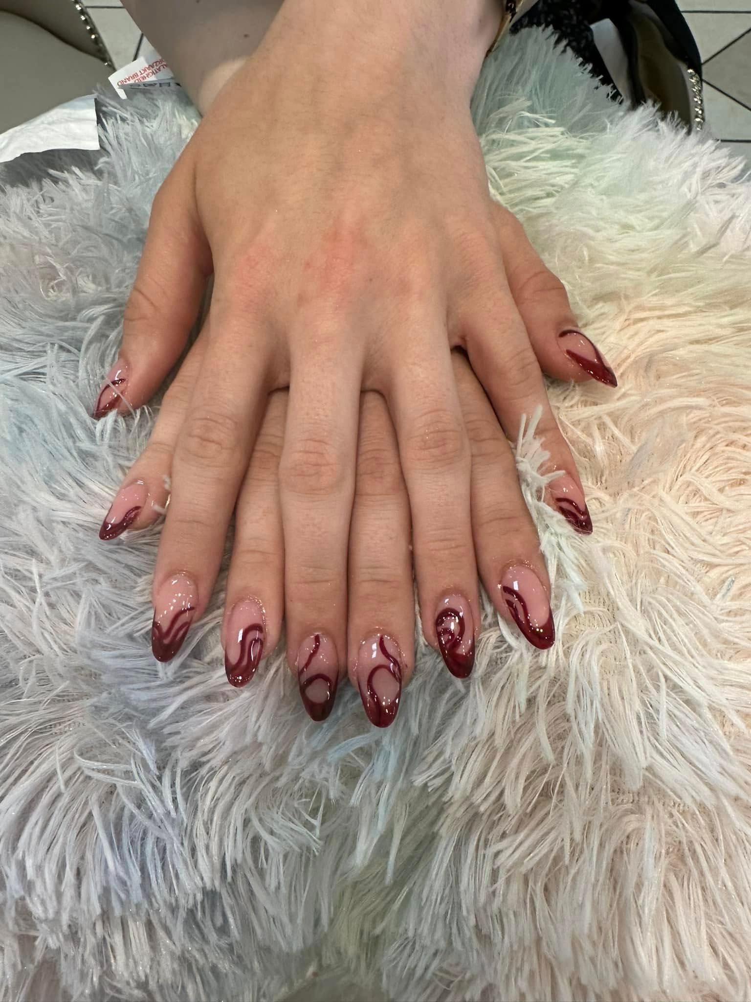 Manicured hands with almond-shaped nails featuring a clear base and dark red swirl designs.