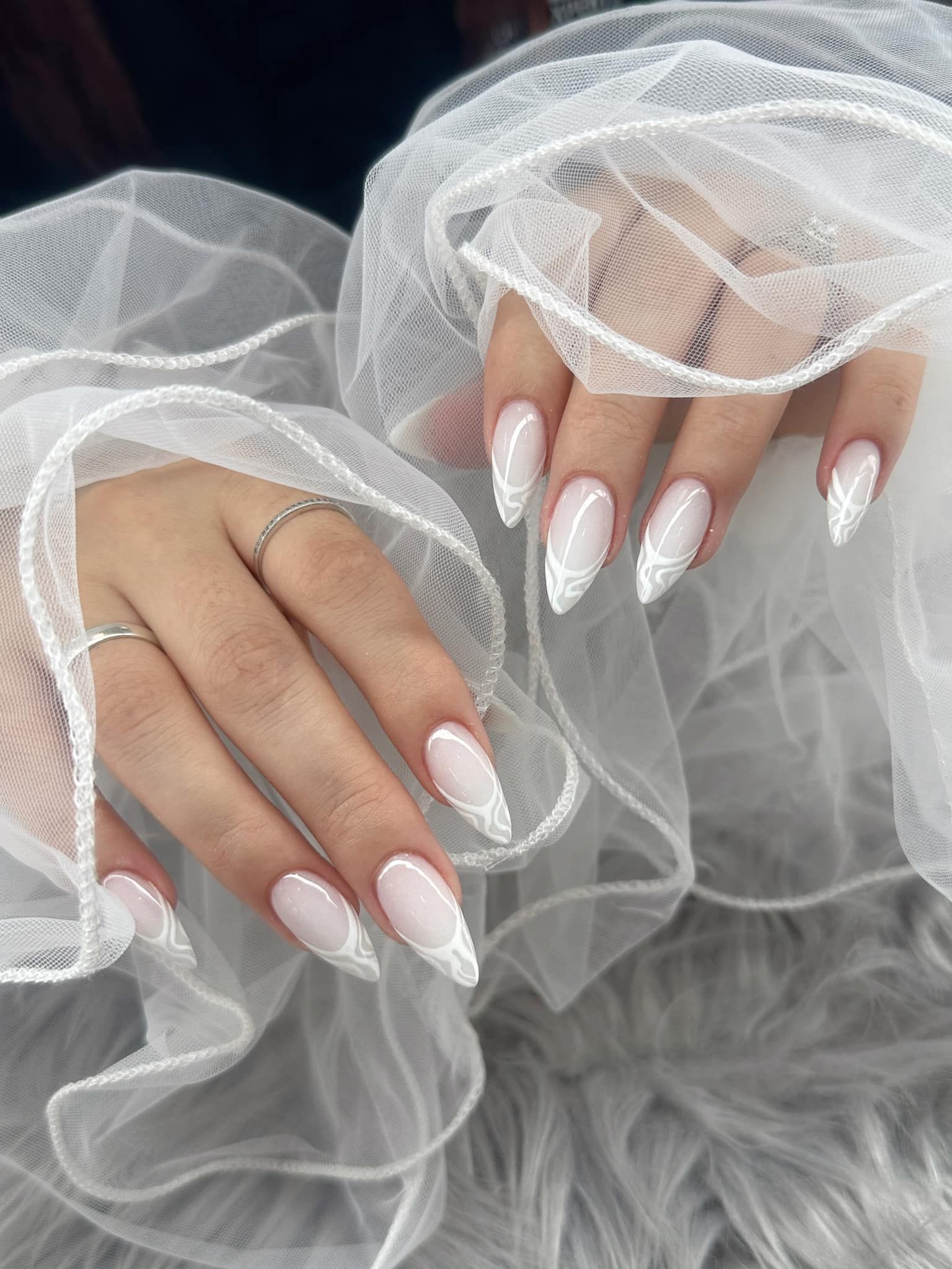 Almond-shaped nails with a white lace-patterned French tip design, held against a soft, white sheer fabric background.