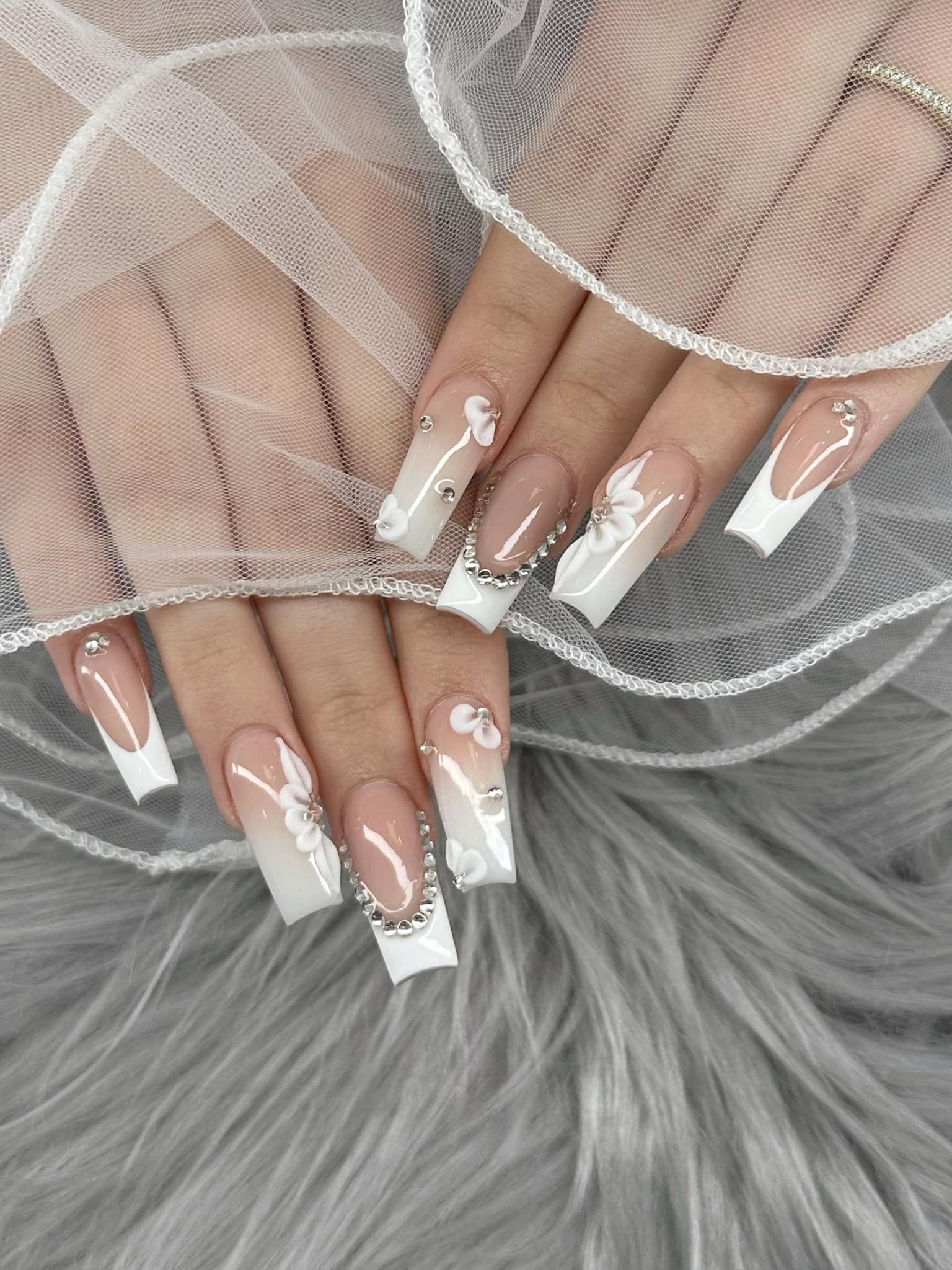Long, square-shaped acrylic nails featuring white floral designs, French tips, and rhinestone accents on a fuzzy background.