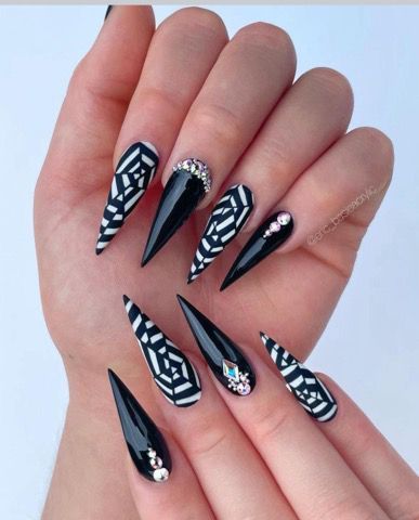 Stiletto-shaped nails with alternating black and white geometric web patterns and solid black polish with crystal accents.