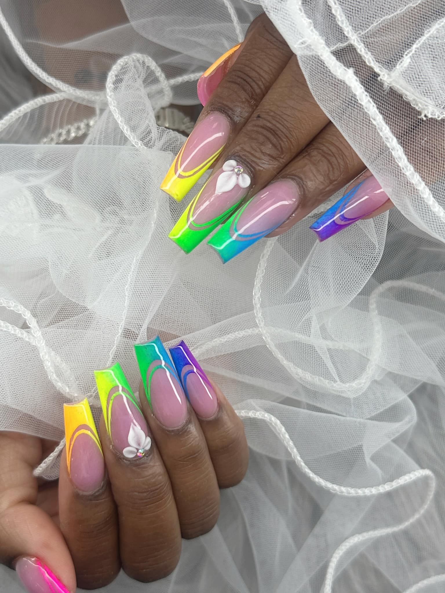 Two hands with long, square-tipped nails featuring a rainbow French manicure, wavy lines, and small white floral accents.