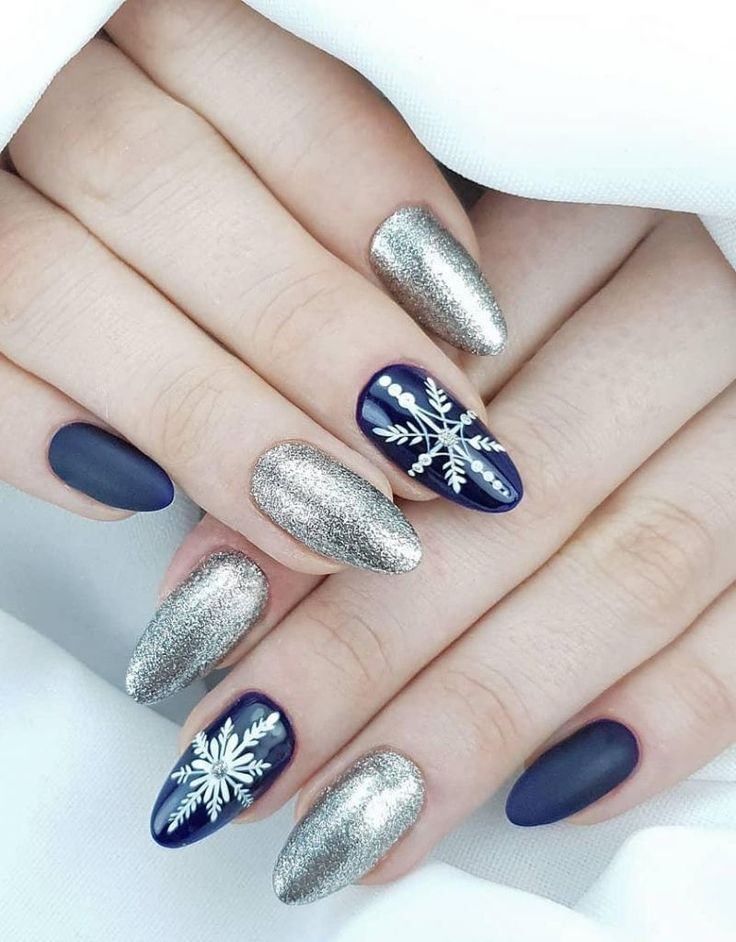 Manicured hands featuring alternating matte navy blue nails and silver glitter nails, some adorned with snowflake art.