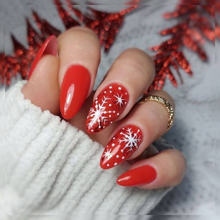 A close-up of almond-shaped nails painted bright red, featuring white snowflake designs and dots against a cozy sweater.