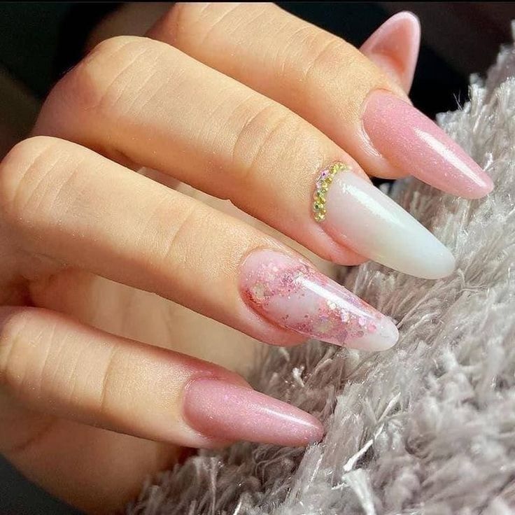 A close-up of a hand with almond-shaped nails featuring pink glitter, a white nail with gold studs, and marble accents.