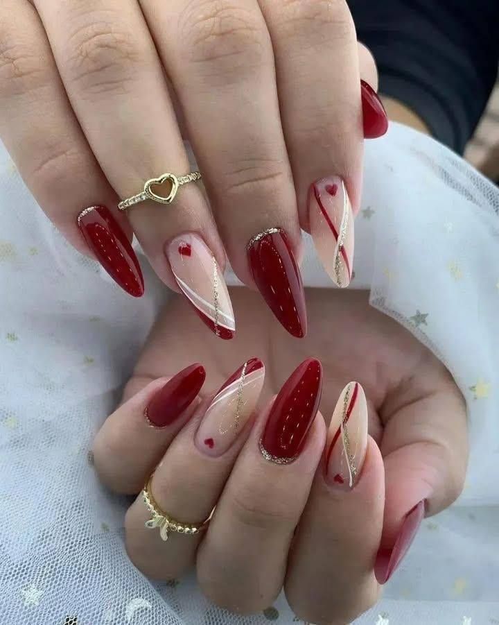 Almond-shaped nails with red polish, sheer accents, heart designs, and gold detailing on both hands wearing gold rings.