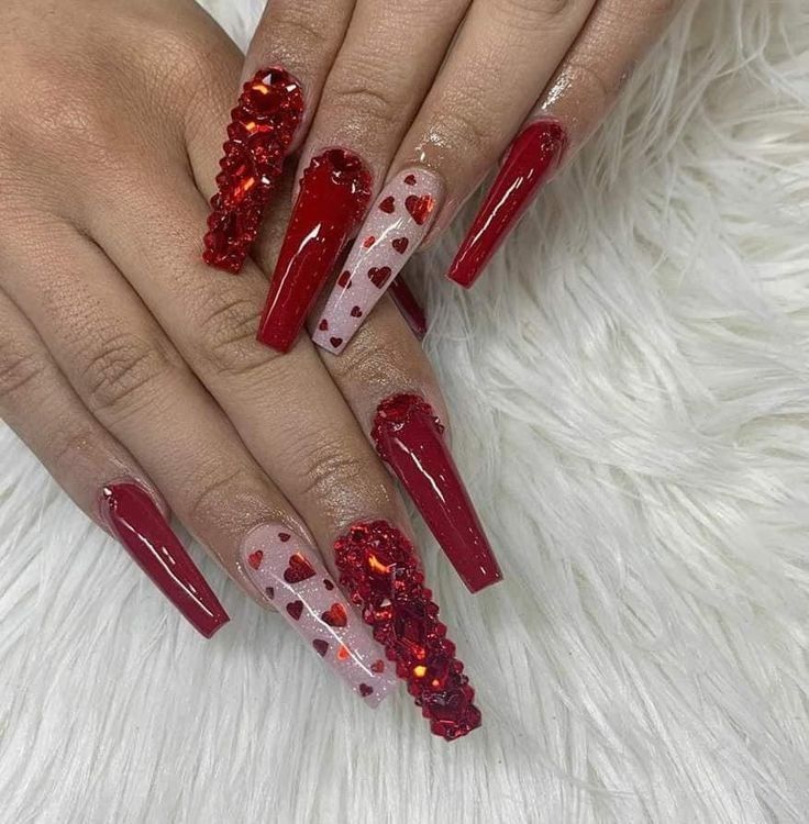 Long coffin-shaped nails painted red, featuring heart patterns, solid red polish, and dense red rhinestone embellishments.