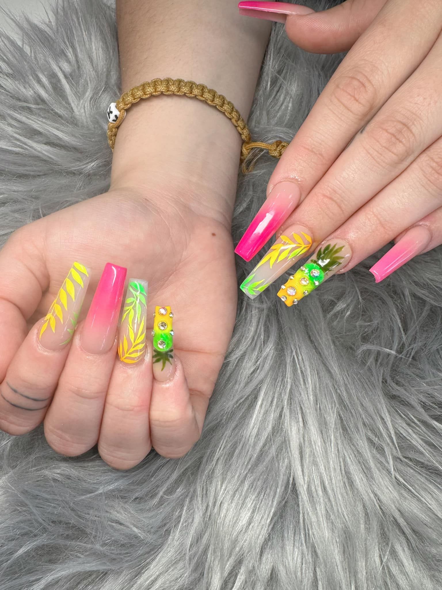 A close-up of hands with long, square-shaped nails featuring bright neon yellow and pink tropical leaf and fruit designs.