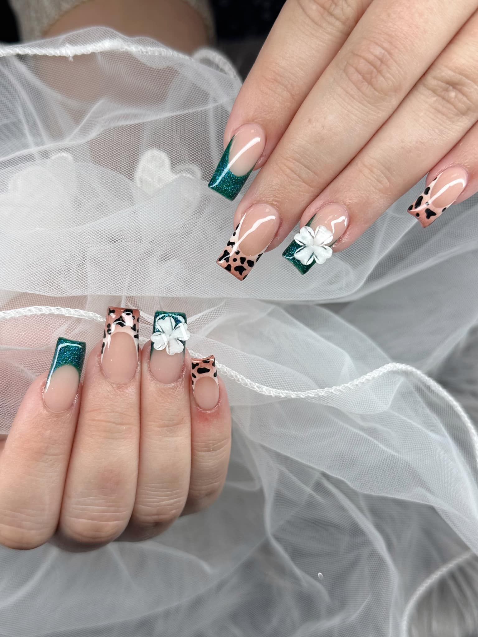 Manicured square nails featuring French tips with dark green glitter, animal print patterns, and white floral embellishments.