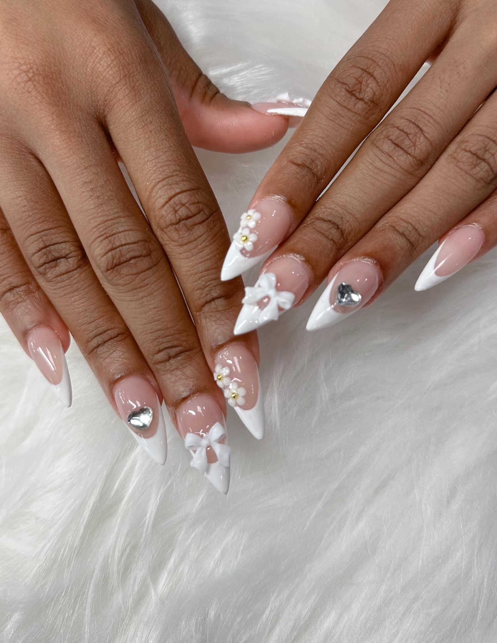 Almond-shaped French tip nails decorated with 3D white bows, delicate floral accents, and small silver heart charms.