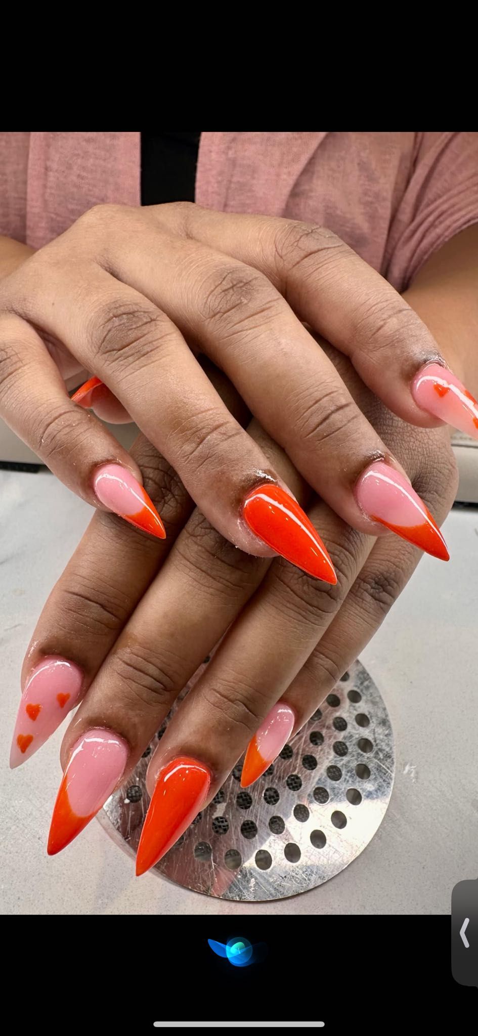 A pair of hands showcasing long, stiletto-shaped nails with a pink base, bright orange tips, and small orange heart accents.