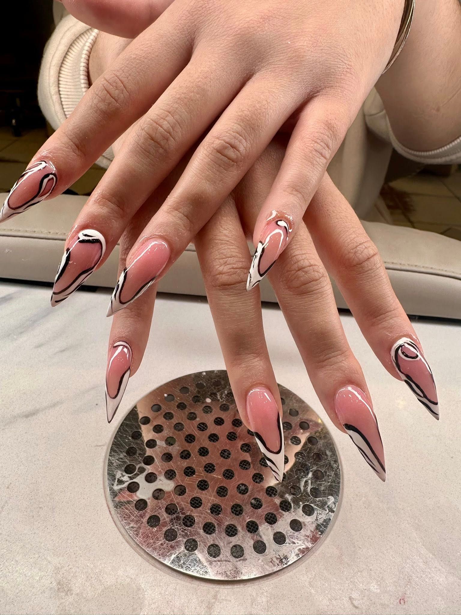 Stiletto-shaped nails with a glossy nude base and bold, black abstract line art accents on a textured surface.