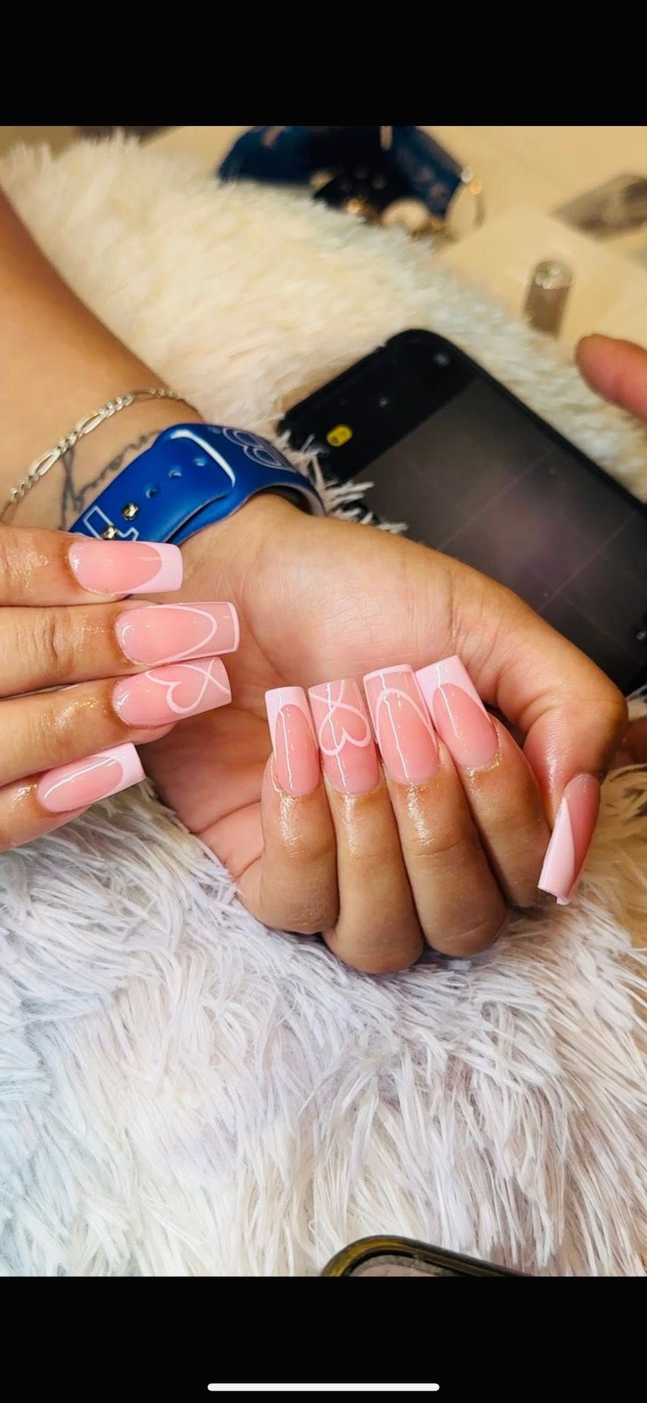 Two hands display long, square-shaped pink acrylic nails with white heart designs on a soft, white textured surface.