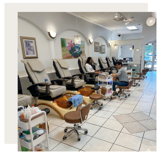 A brightly lit nail salon with rows of pedicure chairs and stools, where a technician tends to a client's feet.