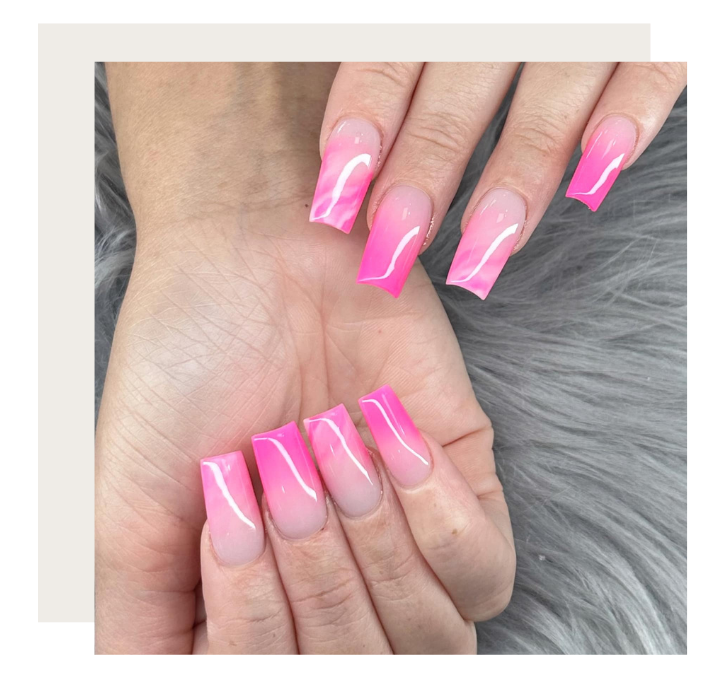 A close-up of hands with square-shaped nails featuring a pink and white ombre design against a grey faux-fur background.