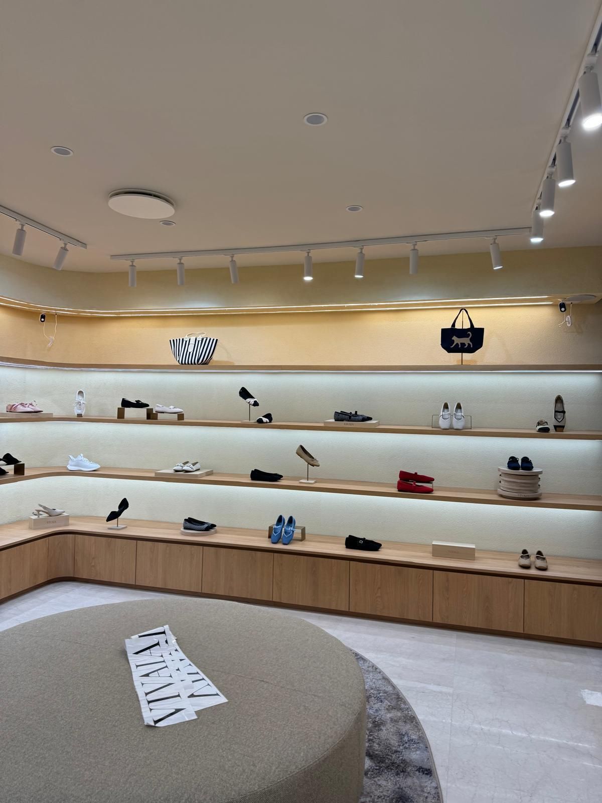 Interno di un negozio di scarpe con scaffali in legno che espongono vari modelli di scarpe e borse. Palette di colori neutri, illuminazione dall'alto.