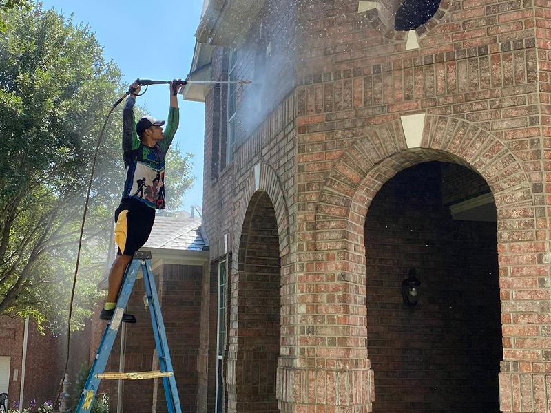 A man is standing on a ladder cleaning a brick building with a pressure washer.