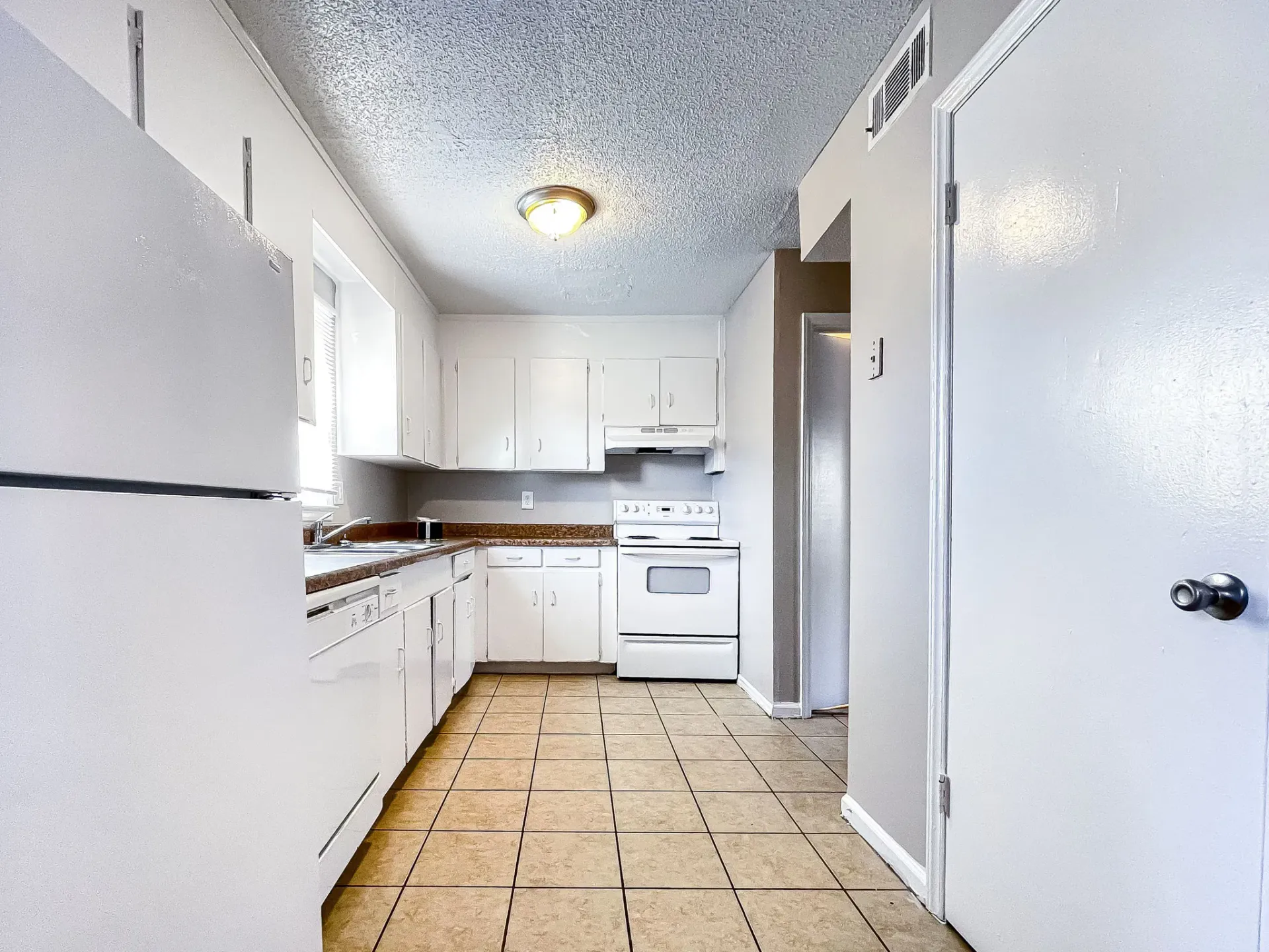Bright white galley kitchen with tiled floor, stove, cabinets, and a windowed sink.