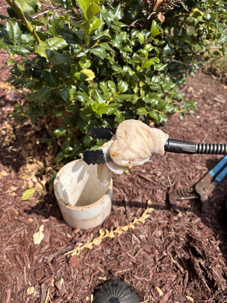 Close-up: a wrapped nozzle in a PVC pipe, possibly for fuel, beside a bush on mulch.