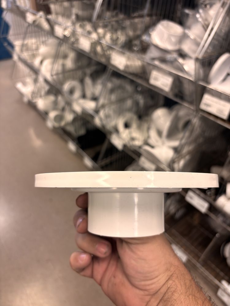 Hand holding a white PVC plumbing fitting with a flat flange and a cylindrical section, inside a store.