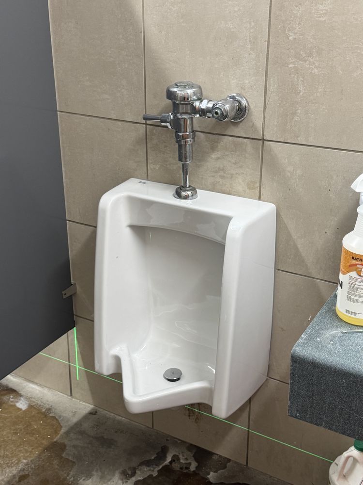 White urinal with chrome flush valve on tiled wall next to gray stall door and cleaning supplies.