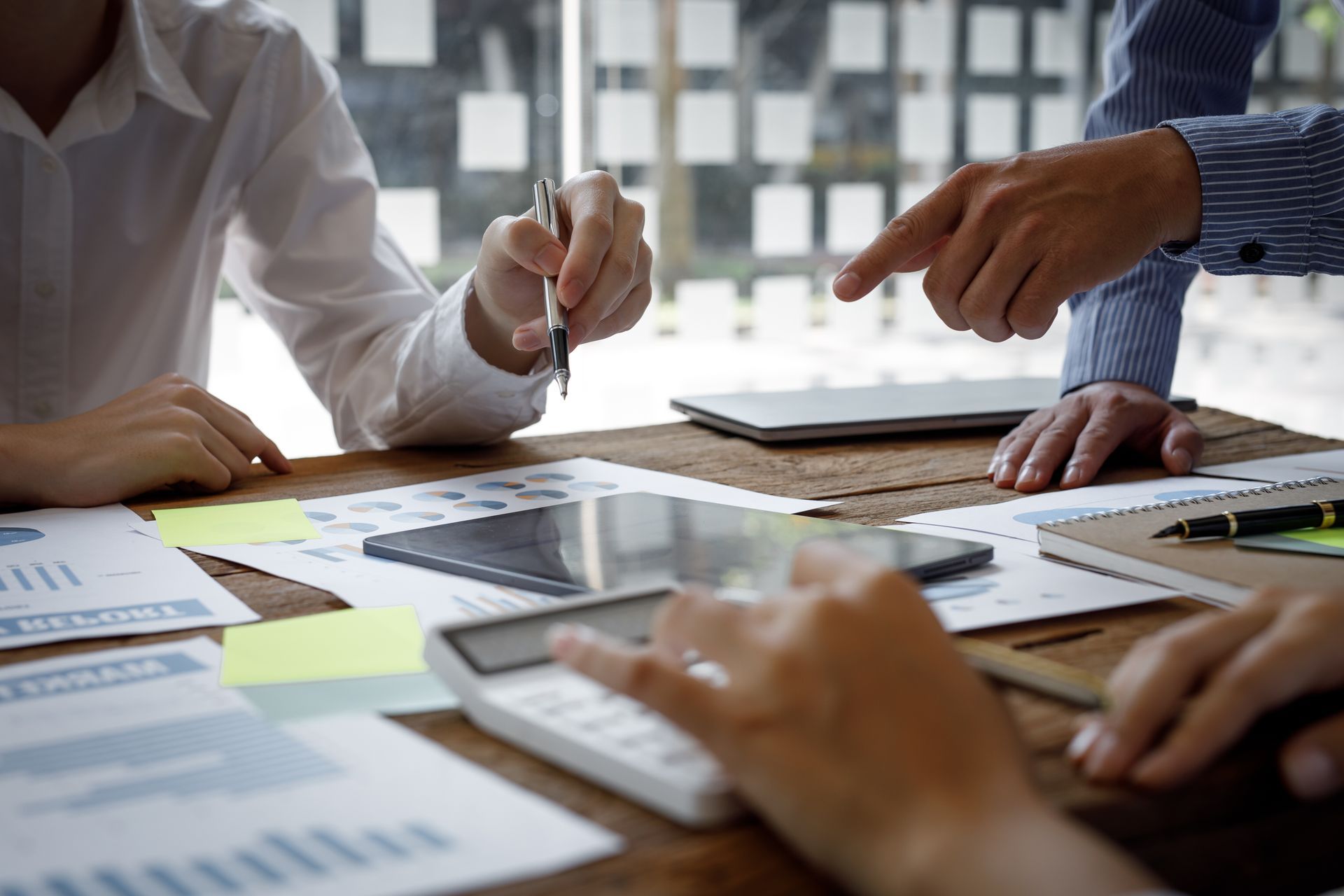 Business meeting at a wooden table with papers, calculator, and laptop; hands pointing and writing.