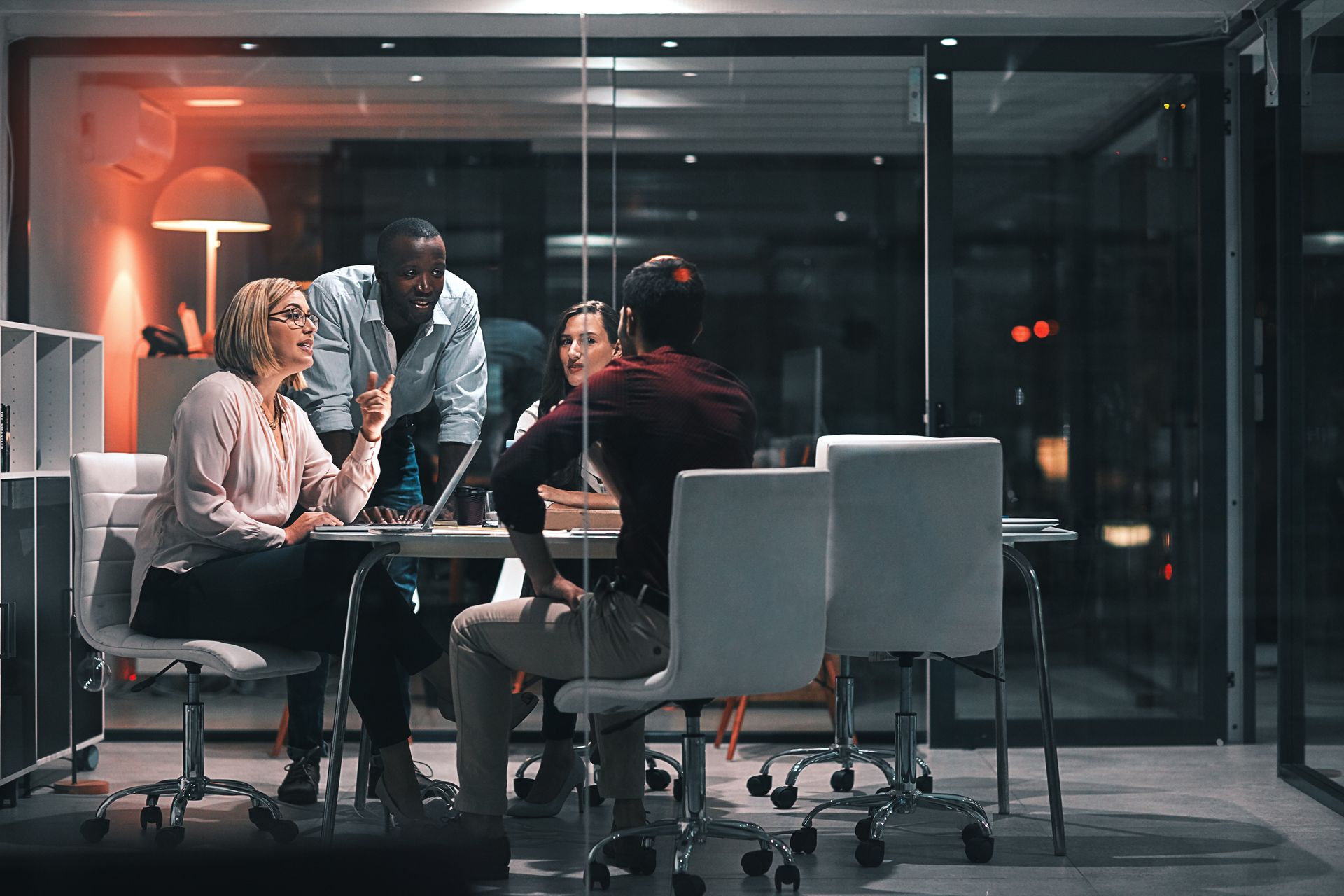 People in a glass-walled office, collaborating around a table at night, illuminated by interior lights.