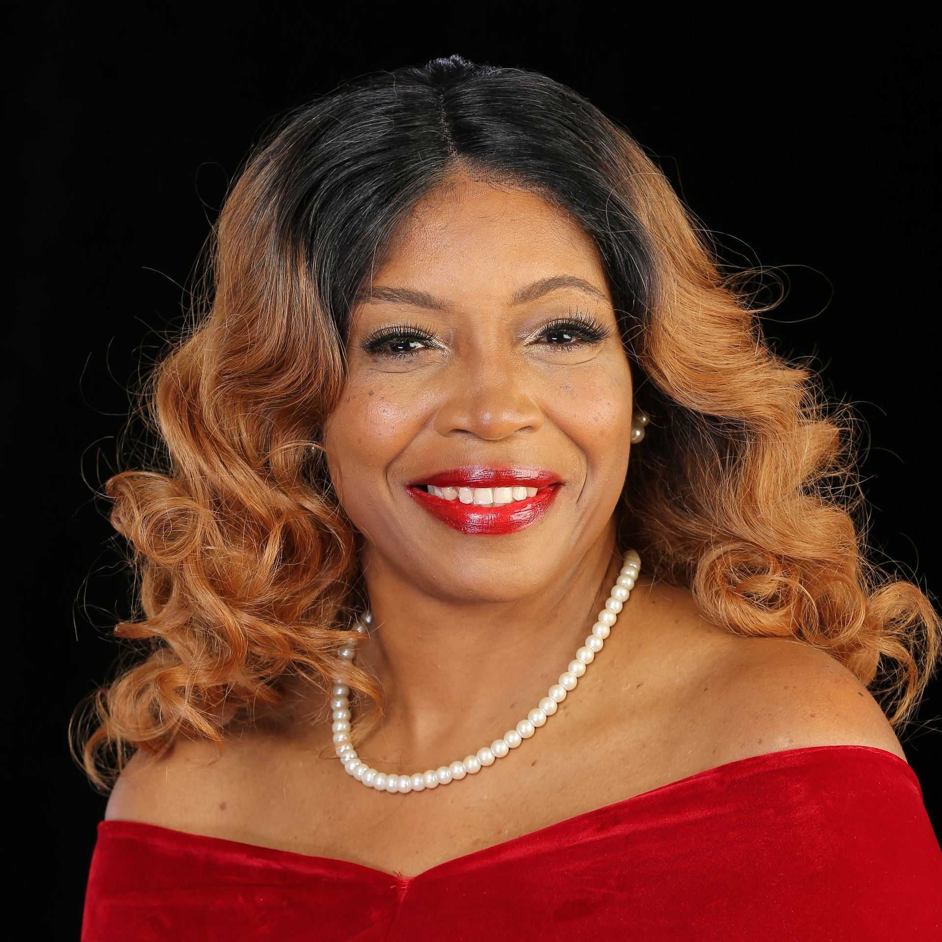 A woman wearing a red dress and a pearl necklace is smiling.