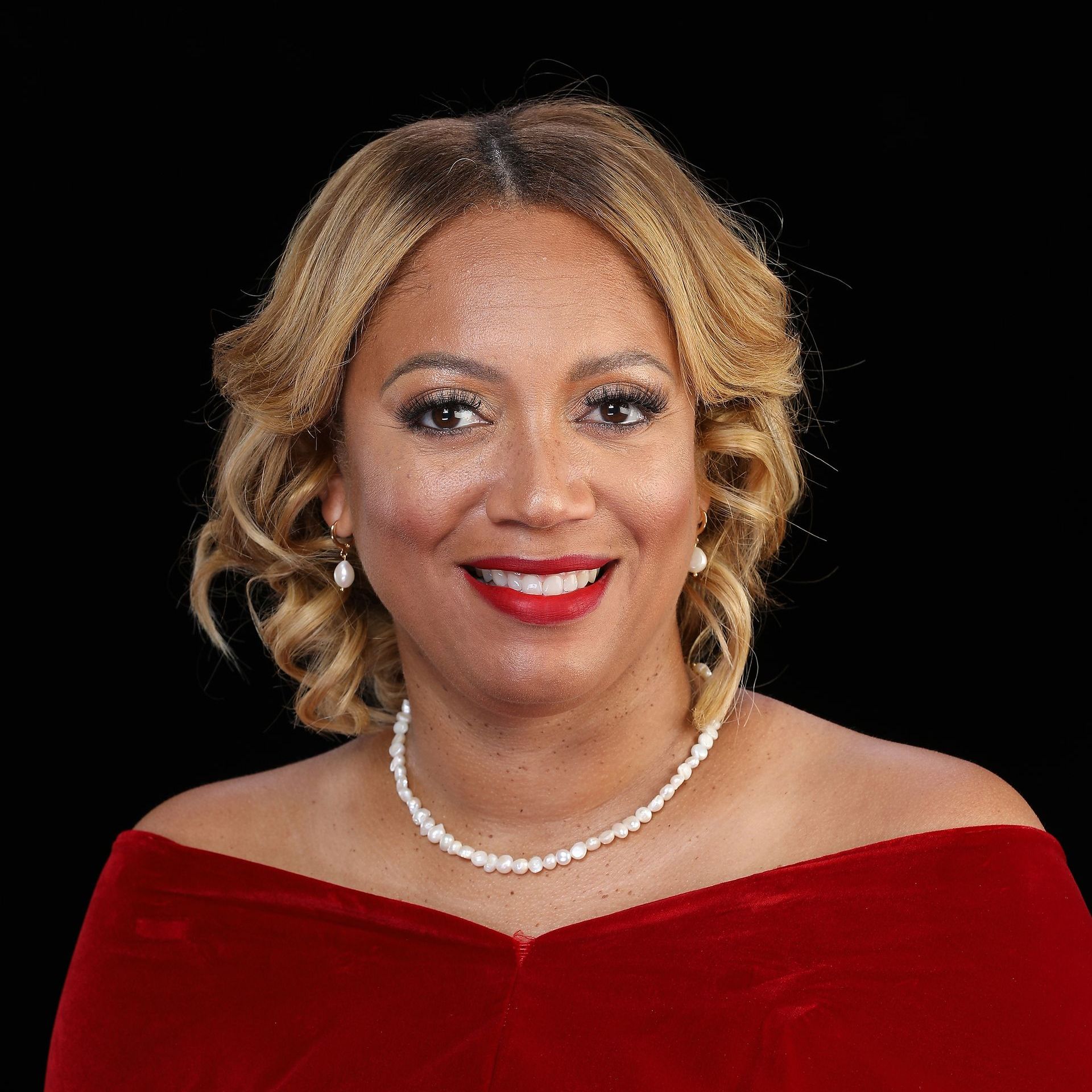 A woman wearing a red dress and a pearl necklace is smiling for the camera.