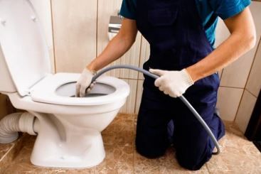Plombier en salopette bleue utilisant un serpent pour déboucher des toilettes blanches dans une salle de bain.