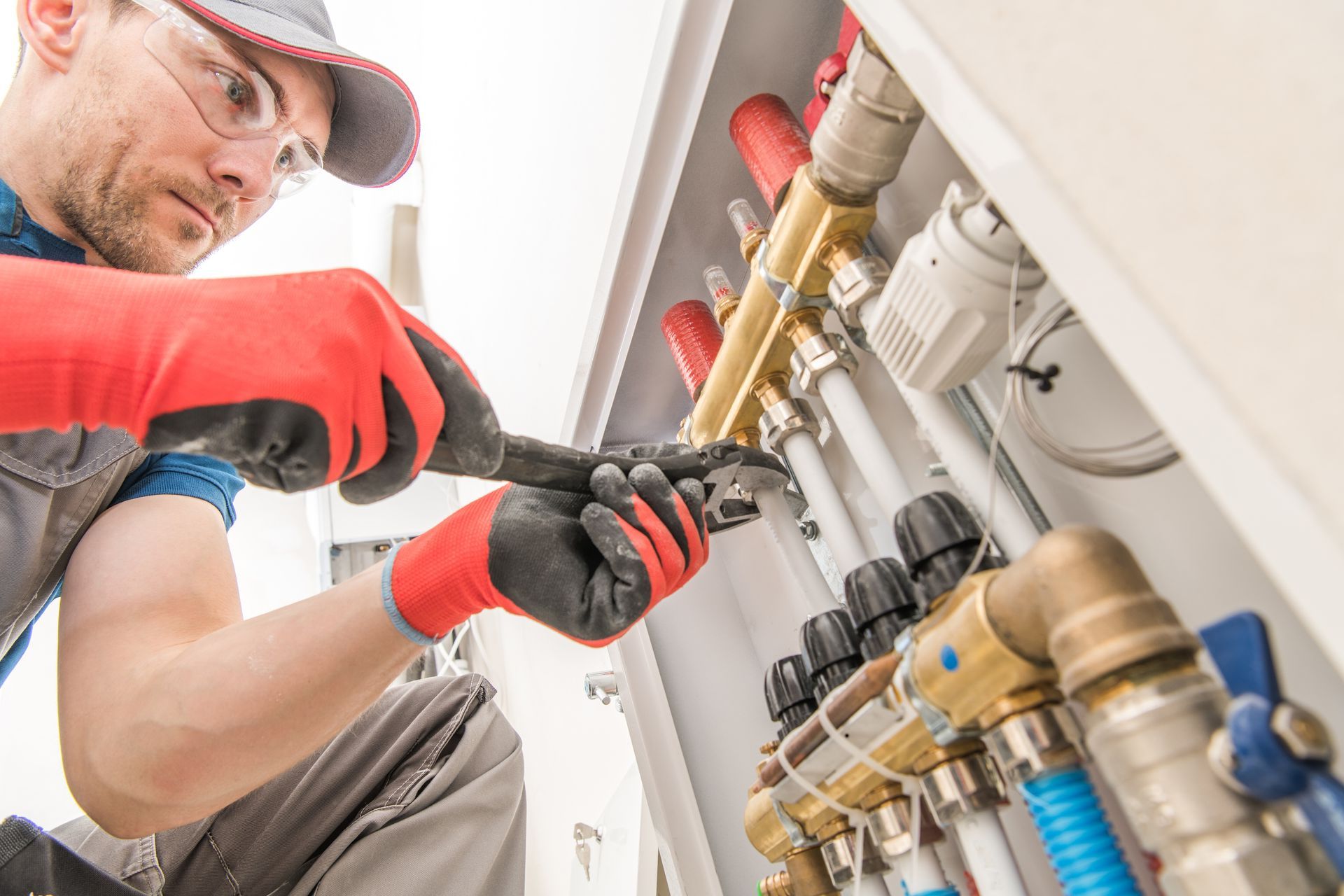 Un plombier portant des lunettes de sécurité et des gants resserre un raccord de tuyau avec une clé à l'intérieur d'une buanderie.