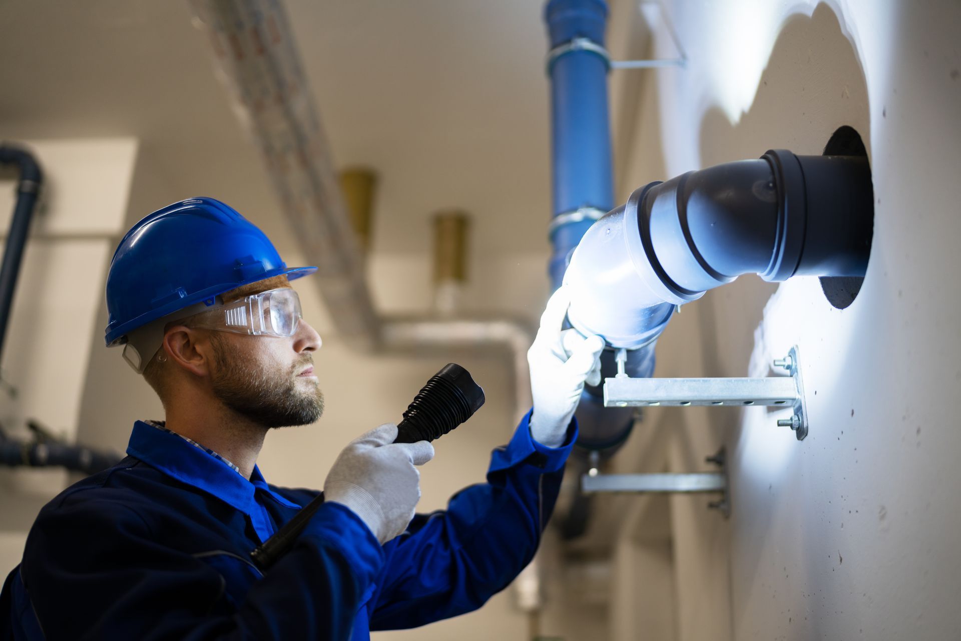Un homme en tenue de travail bleue inspecte des tuyaux avec une lampe de poche, portant des lunettes de sécurité et un casque.