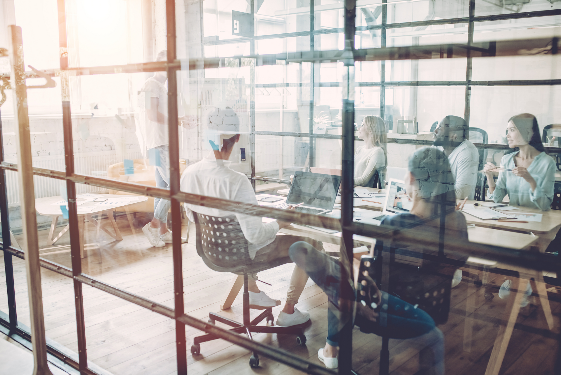People in a meeting room, behind glass; working at tables, discussing and gesturing; bright lighting.