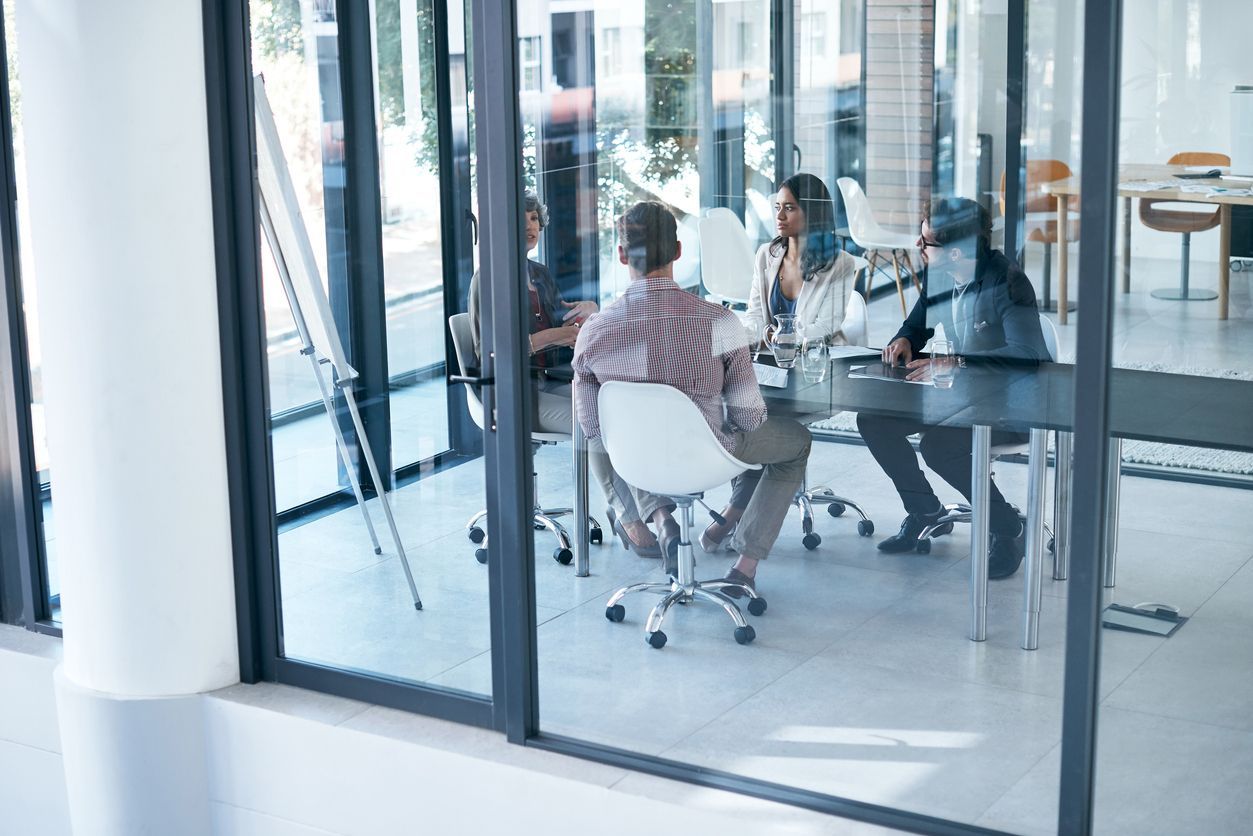 People in a meeting room, sitting around a table, brainstorming. Glass wall reflects outside.