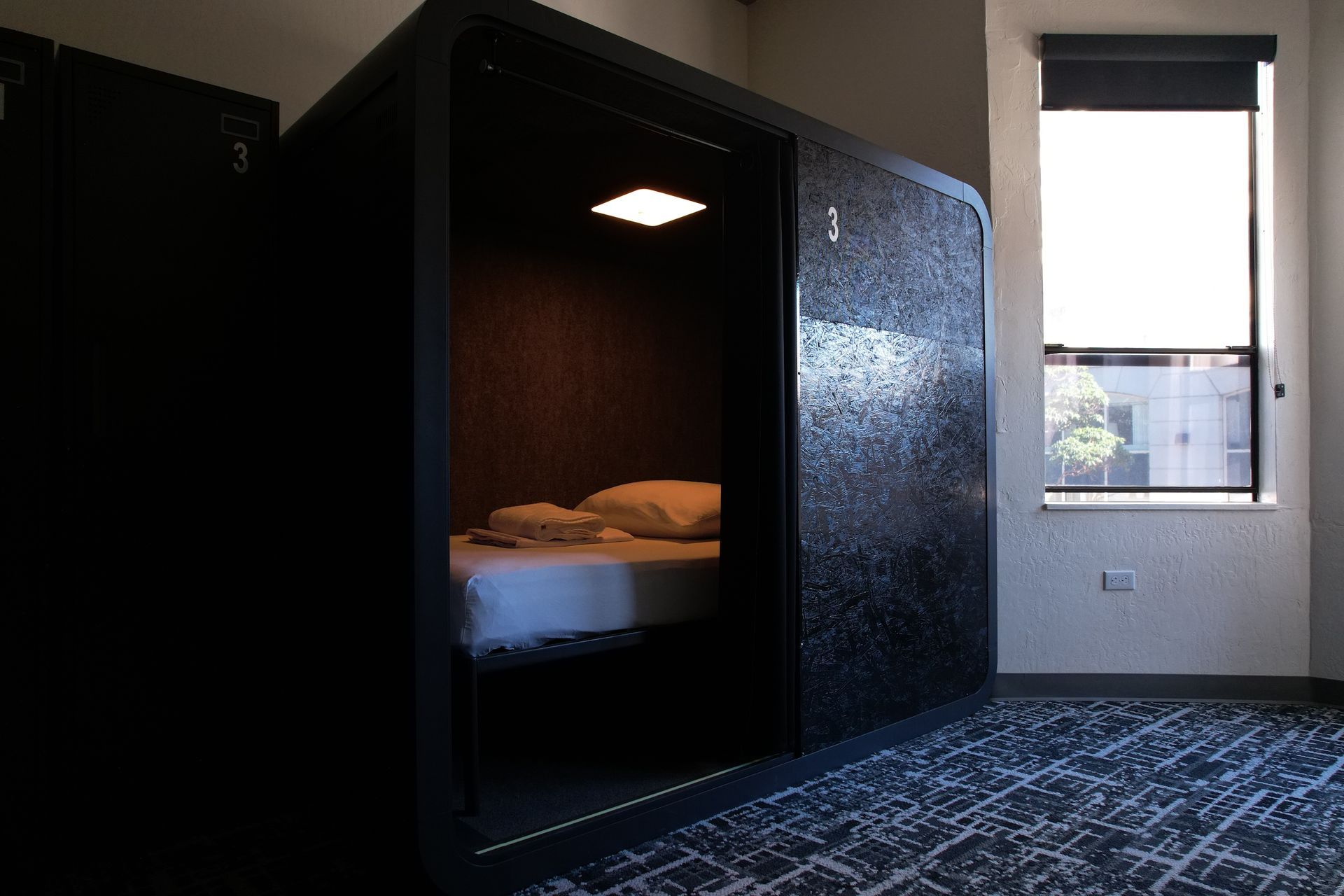 Capsule hotel room with a bed inside, next to a window with a rolled-up blind.
