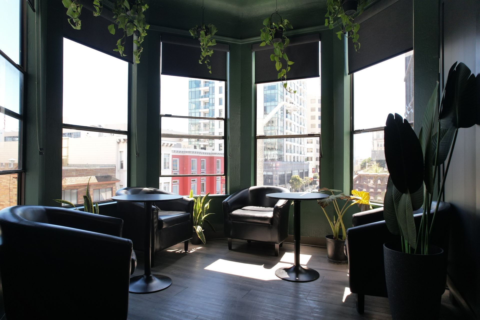 Lounge area with black chairs and small tables by large windows overlooking a city. Green walls and hanging plants.