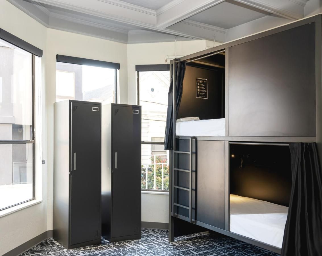 Woman in a black pod room using a phone, sitting on a bed. Pod door is open.