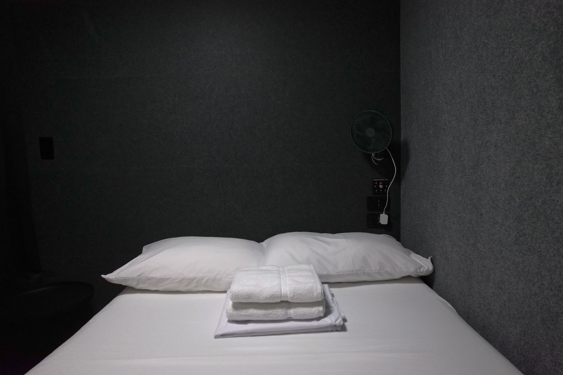 Bed with white linens, two pillows, folded towels, against dark gray walls.