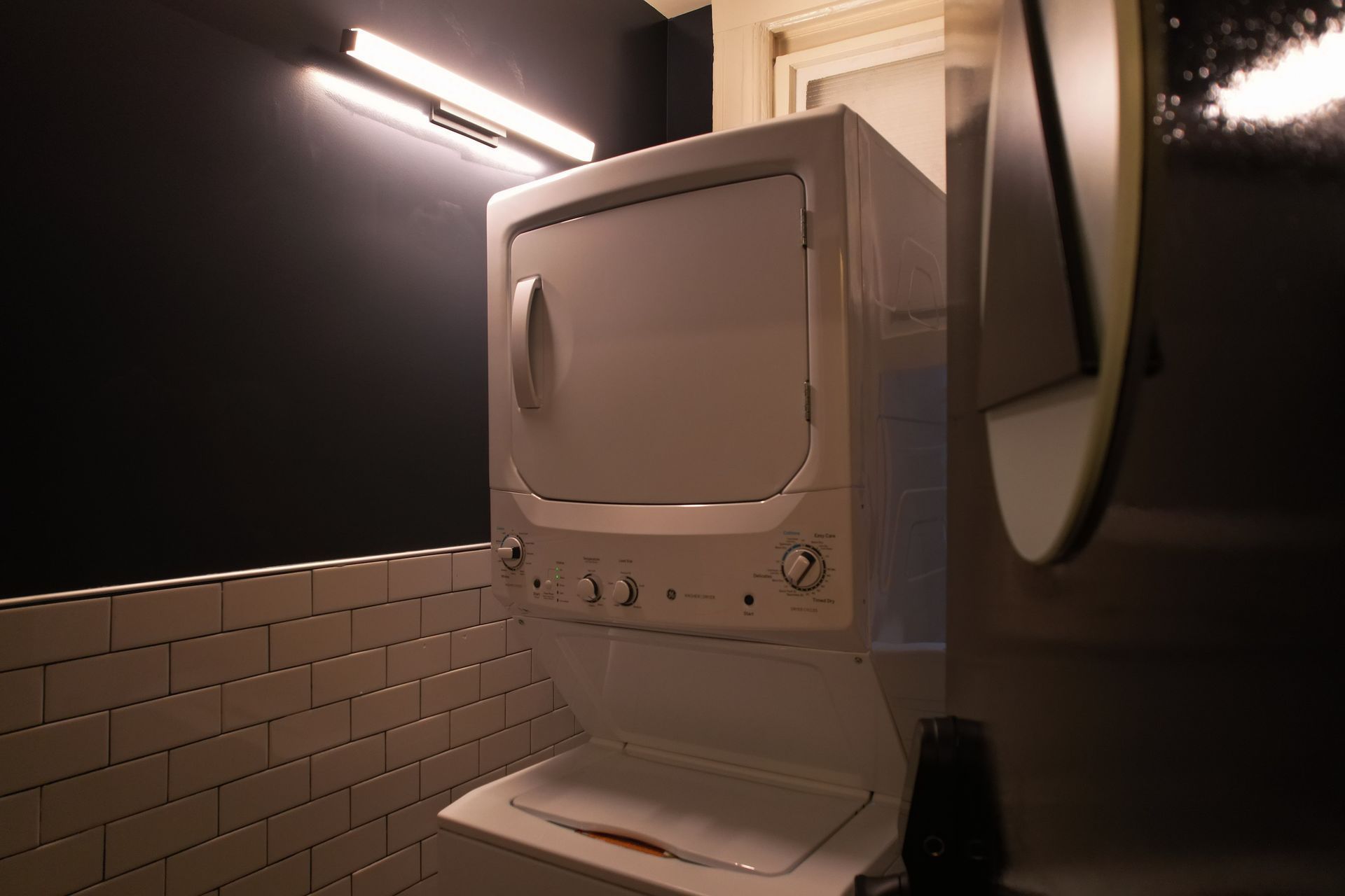 Stackable white washer and dryer in a small bathroom with black and white tiled walls and a round mirror.