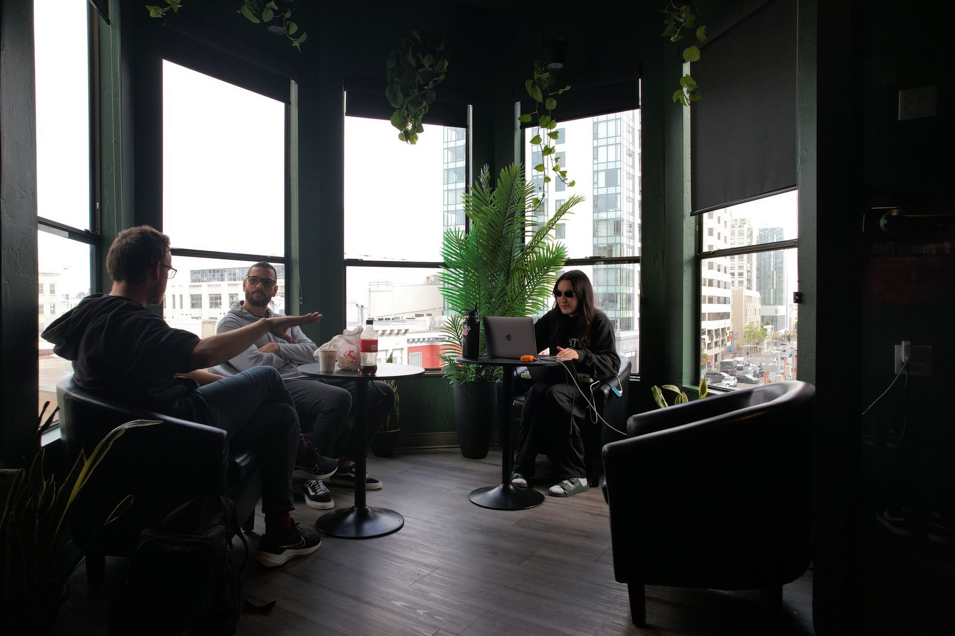 Three people in a dark room with large windows, working or conversing, two seated at a table, one at a laptop.