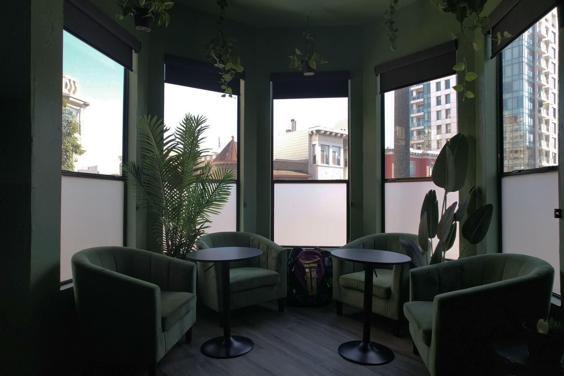 Three people in a dark room with large windows, one using a laptop, conversing at tables, greenery and city view.