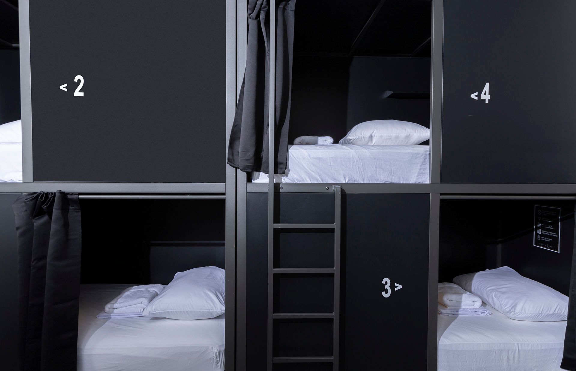Black and white capsule hotel rooms, numbered 2, 3, and 4, each with a bed, pillow, and privacy curtain.