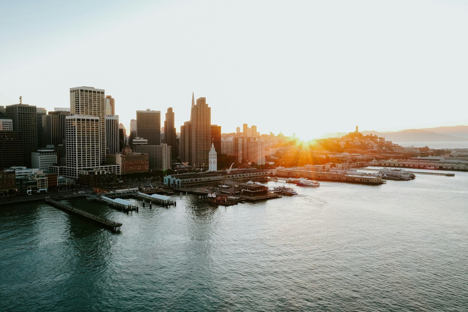 City skyline at sunset, with buildings lining the waterfront. Sun glows brightly, reflecting on the water.