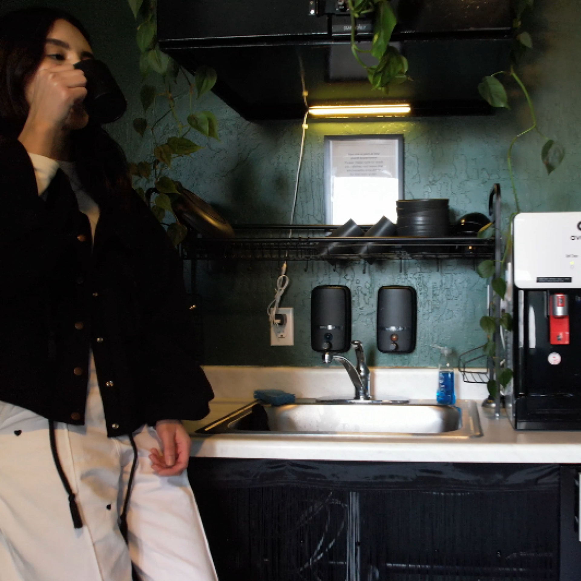 Person drinking from a mug in a small kitchen. Plants, sink, water cooler, black jacket, white pants.