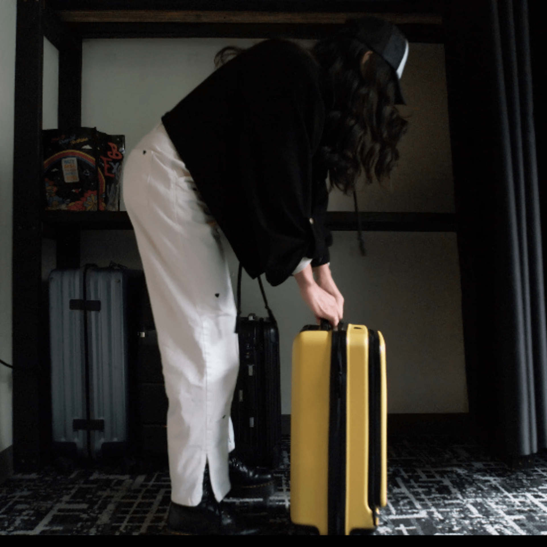 Person in black hoodie and white pants, reaching for a yellow suitcase in a room.
