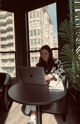 Woman working on laptop at a dark table near a window.  Tall buildings and plant visible.