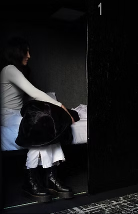 Woman seated in a dark, narrow room, next to a bed, holding a dark jacket. Room labeled 
