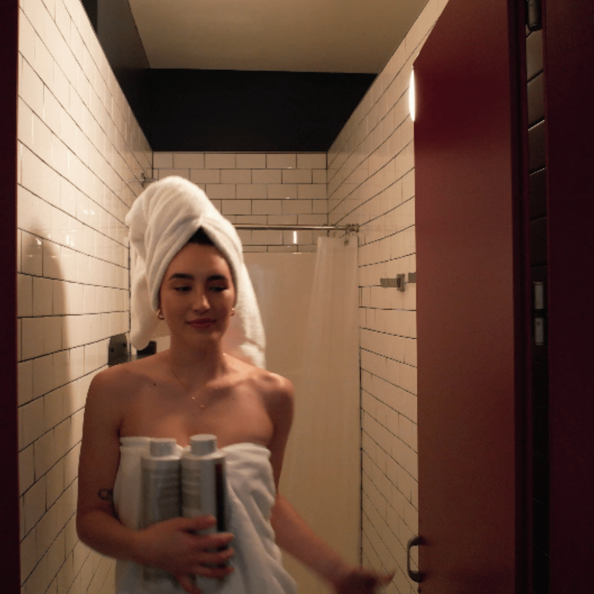 Woman wrapped in a towel exits a small shower, holding two shampoo bottles.