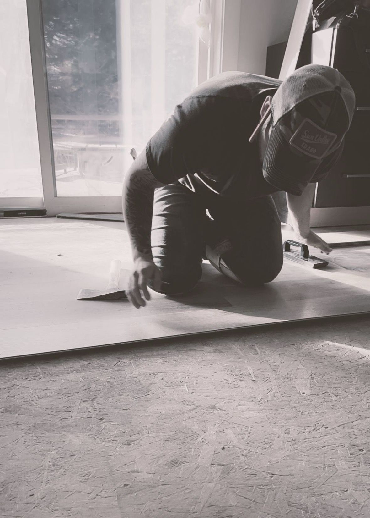 Man kneeling, cutting material on the floor indoors; wearing a cap.