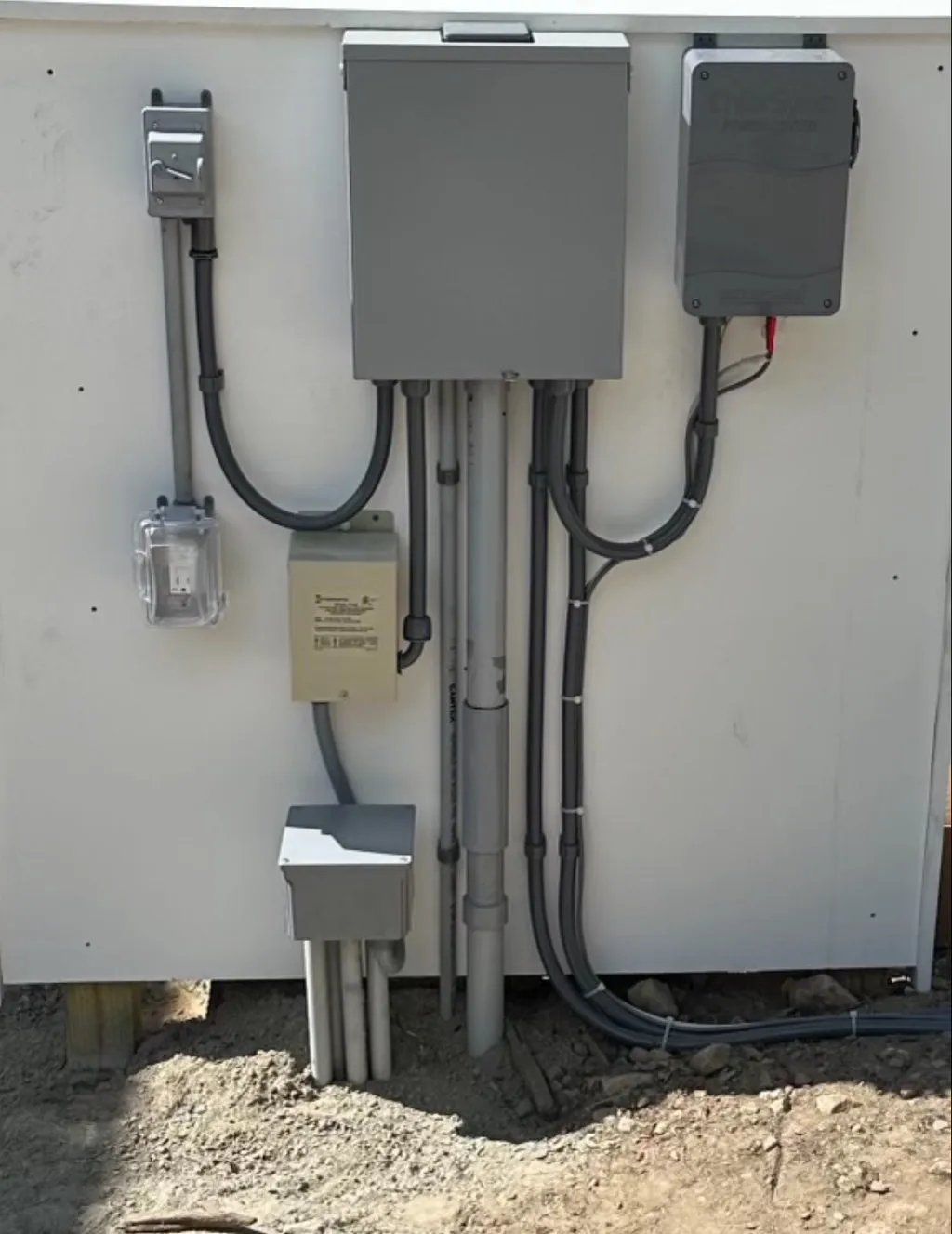 Electrical boxes and conduits mounted on a white wall, outdoors on gravel.