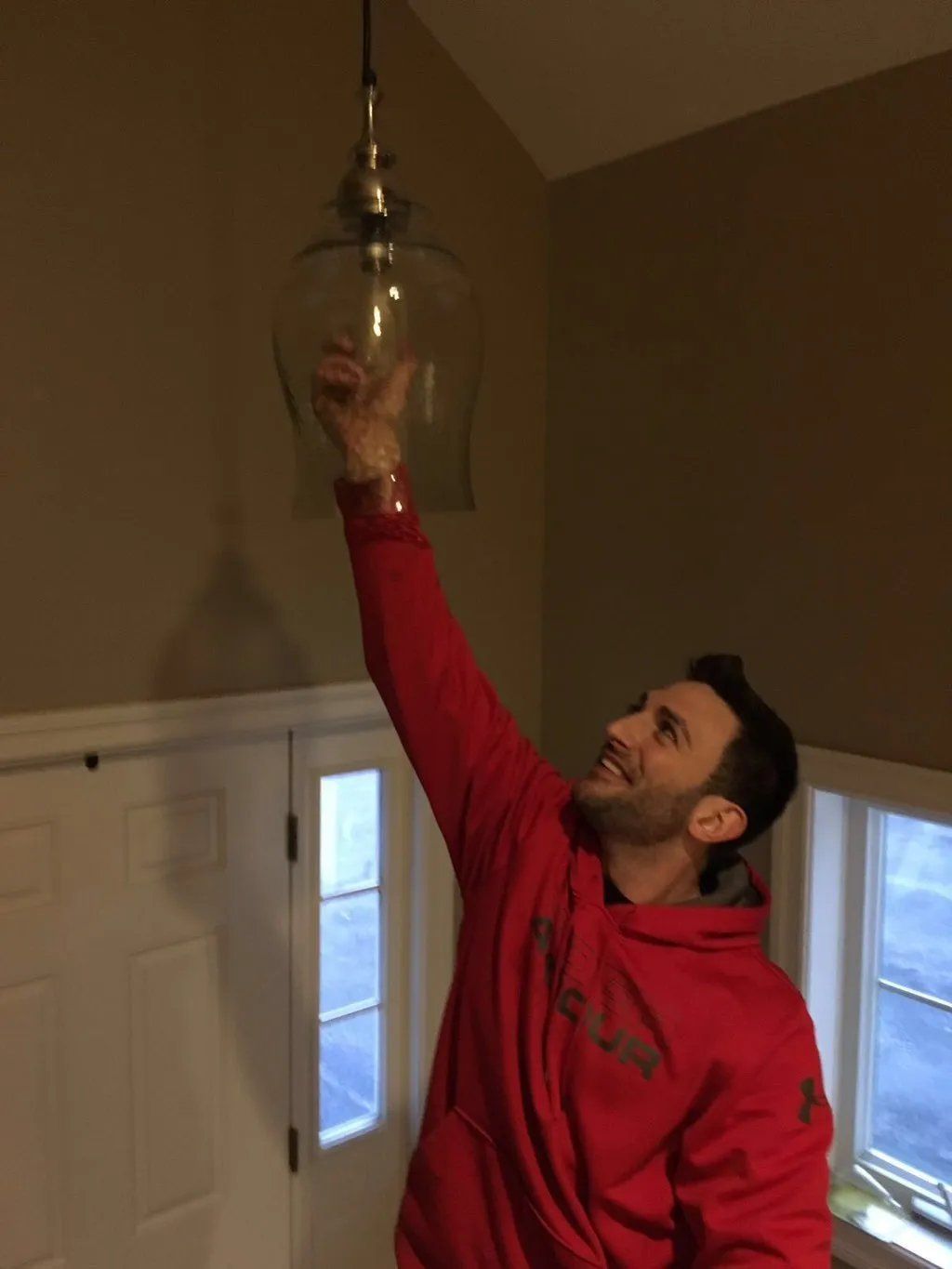 Man in red hoodie reaching up toward glass pendant light. Smiling.